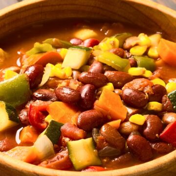 A close-up of a hearty bowl of spicy vegetarian chili filled with kidney beans, black beans, corn, zucchini, bell peppers, carrots, and tomatoes in a rich, savory broth. The vibrant mix of vegetables and beans sits in a rustic wooden bowl, showcasing a colorful and comforting plant-based meal.