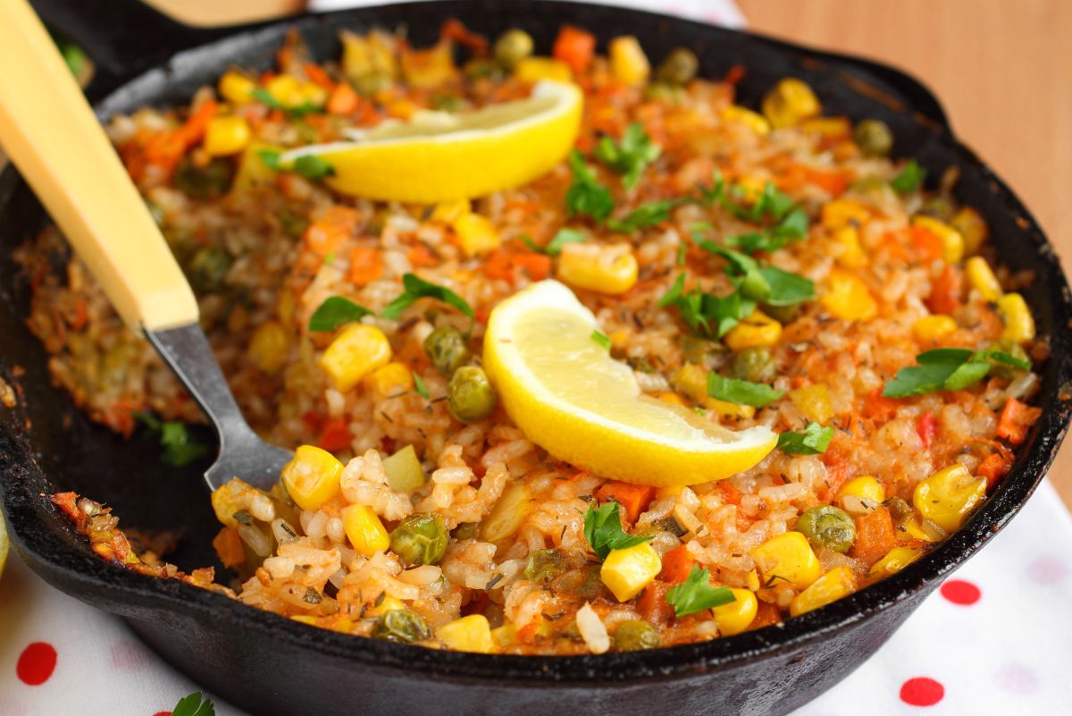 A cast iron skillet filled with taco beef and rice skillet dinner, topped with corn, peas, diced peppers, fresh parsley, and lemon wedges for garnish. A serving spoon rests inside, ready to scoop the colorful, hearty meal.