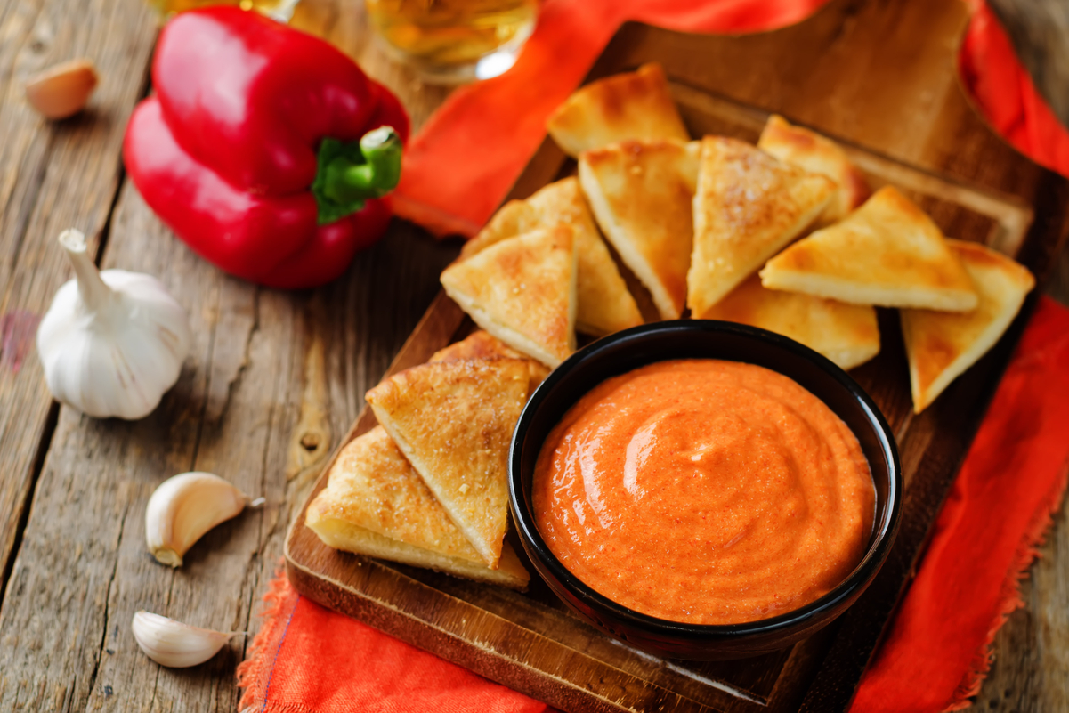 Creamy red pepper dip in a black bowl, served on a wooden board with golden toasted pita triangles. Surrounding the board are fresh red bell peppers, whole garlic bulbs, and cloves on a rustic wooden table with a bright orange cloth for a warm, inviting presentation.
