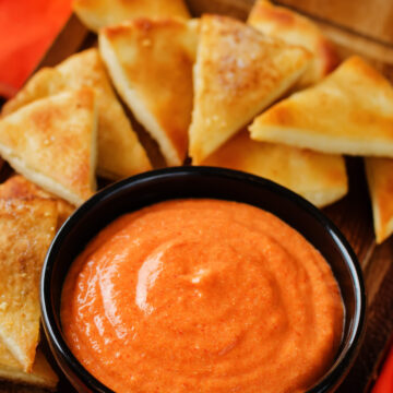 A black bowl filled with smooth, creamy red pepper dip, its vibrant orange-red color glistening under the light, served on a wooden board with golden, toasted pita bread triangles arranged in the background.