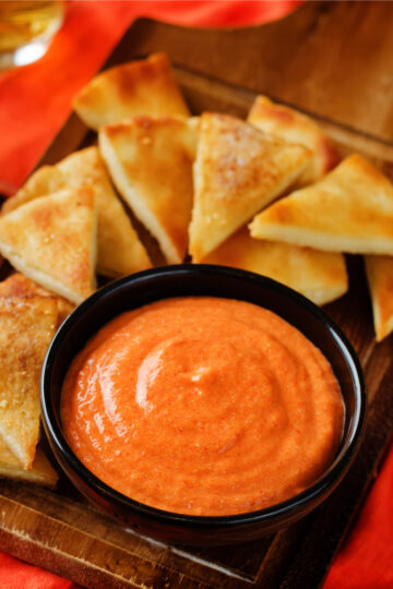 A black bowl filled with smooth, creamy red pepper dip, its vibrant orange-red color glistening under the light, served on a wooden board with golden, toasted pita bread triangles arranged in the background.