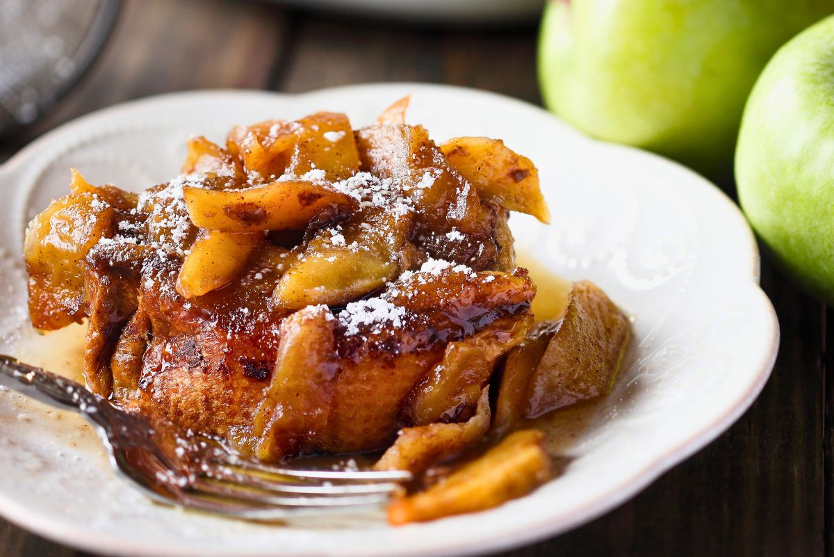 Slow cooker apple cinnamon French toast casserole served on a white plate, topped with tender caramelized apples, warm cinnamon sauce, and a light dusting of powdered sugar. Fresh green apples in the background add a cozy autumn touch.