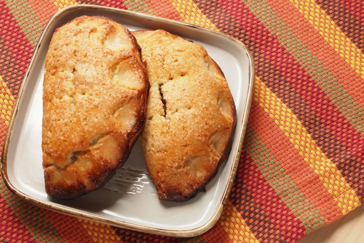 Two golden spiced maple-pumpkin hand pies with a crisp, sugared crust rest on a white dish. The flaky pastries are baked to a rich brown and set on a colorful woven fall placemat with orange, red, yellow, and green tones.