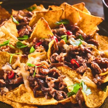 Plate of beef and bean nachos: crispy tortilla chips topped with seasoned ground beef, melted cheese, parsley, and sliced red chile peppers on a dark platter, warm and melty. Keywords: beef and bean nachos, easy appetizer, game day snack, weeknight dinner.