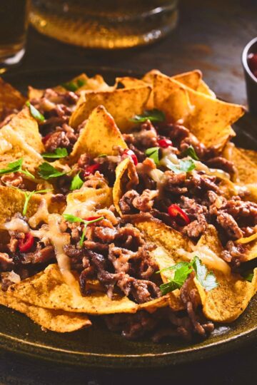 Plate of beef and bean nachos: crispy tortilla chips topped with seasoned ground beef, melted cheese, parsley, and sliced red chile peppers on a dark platter, warm and melty. Keywords: beef and bean nachos, easy appetizer, game day snack, weeknight dinner.