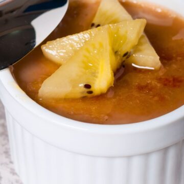 A white ramekin filled with rich coffee caramel baked custard, topped with golden kiwi slices. A spoon rests on the edge, ready to dig into the smooth custard with its glossy caramelized surface.