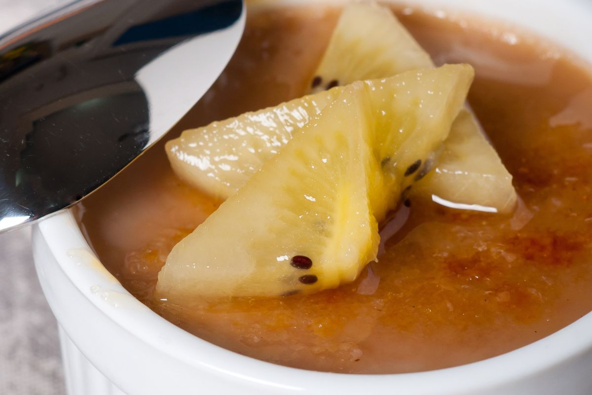 Close-up of a creamy coffee caramel baked custard in a white ramekin, topped with fresh golden kiwi slices. A shiny spoon rests on the edge, highlighting the custard’s smooth caramelized surface and glossy fruit garnish.
