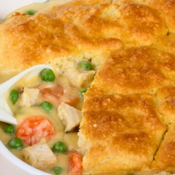 Golden biscuit crust tops a creamy chicken potpie in a white baking dish. The filling is rich with chunks of chicken, carrots, and green peas in savory gravy, peeking through where a portion has been scooped out. The dish shows a contrast of flaky, browned topping and hearty, colorful vegetables in sauce.
