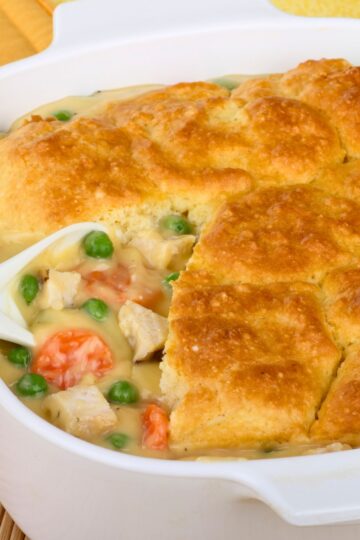 Golden biscuit crust tops a creamy chicken potpie in a white baking dish. The filling is rich with chunks of chicken, carrots, and green peas in savory gravy, peeking through where a portion has been scooped out. The dish shows a contrast of flaky, browned topping and hearty, colorful vegetables in sauce.