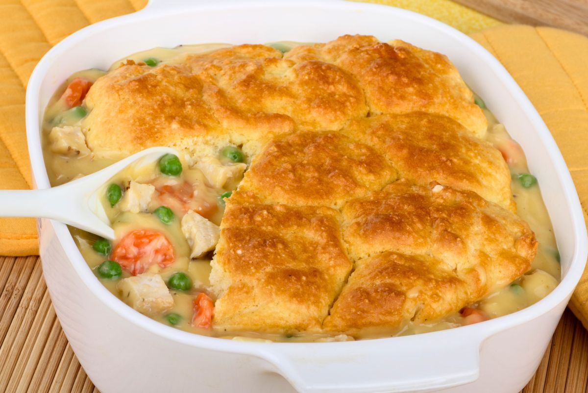 Baked chicken potpie in a white casserole dish topped with golden biscuit crust. A serving spoon reveals the creamy filling made of tender chicken, carrots, and green peas in rich gravy beneath the flaky topping. The dish rests on a wooden surface with a yellow oven mitt nearby.