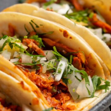 Soft tortillas filled with shredded adobo pork, topped with fresh chopped onions and cilantro. The tacos are arranged side by side on a dark serving tray, showing the rich red pork filling and fresh green herbs for a vibrant Mexican dinner presentation.