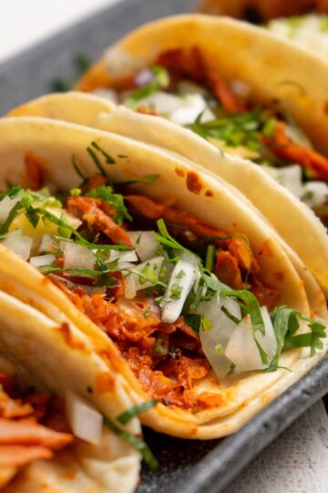 Soft tortillas filled with shredded adobo pork, topped with fresh chopped onions and cilantro. The tacos are arranged side by side on a dark serving tray, showing the rich red pork filling and fresh green herbs for a vibrant Mexican dinner presentation.