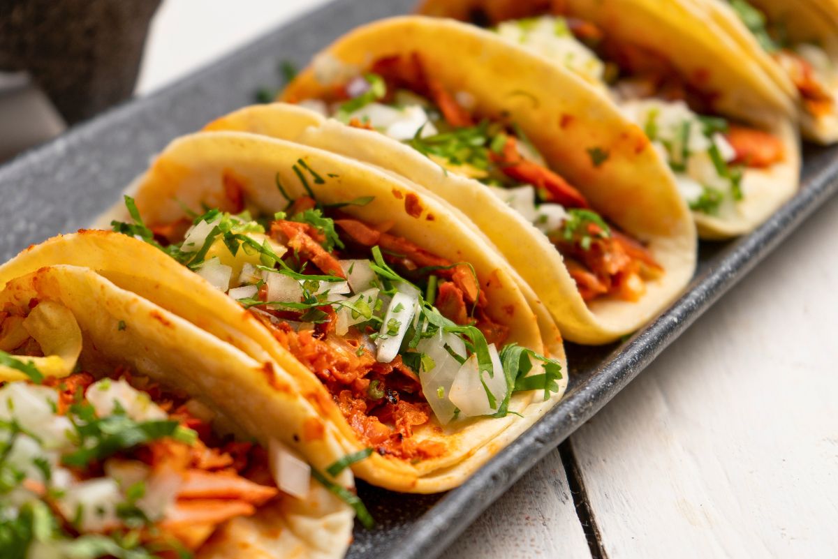 A row of soft corn tortillas filled with shredded adobo pork, topped with chopped white onions and fresh cilantro. The tacos are neatly arranged on a rectangular dark serving tray, showing the vibrant red pork filling and green garnish against the warm golden tortillas.