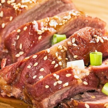 Close-up of juicy teriyaki pork tenderloin slices on a wooden board, topped with toasted sesame seeds and fresh green onions. The glossy glaze highlights the tender texture, creating a savory and mouthwatering presentation.