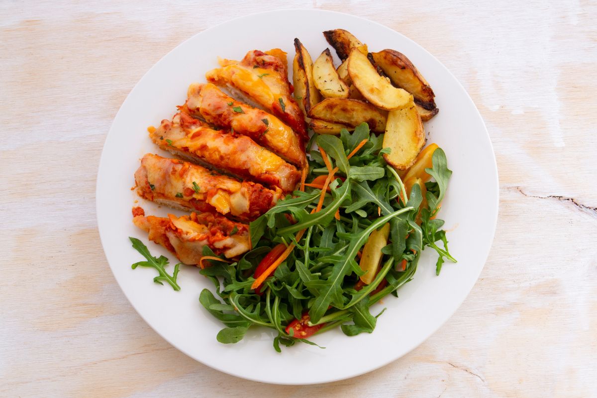 Sliced baked chicken parmesan topped with melted cheese and tomato sauce, served with golden roasted potato wedges and a fresh arugula salad with shredded carrots and red bell pepper on a white plate.