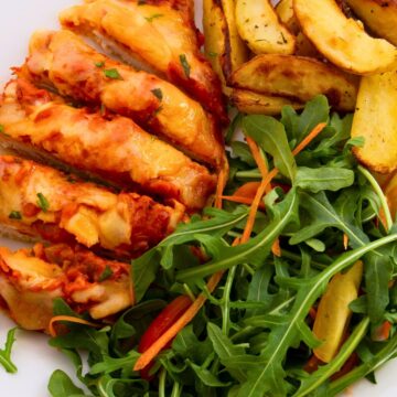 Close-up of sliced baked chicken parmesan topped with melted cheese and tomato sauce, served with golden roasted potato wedges and a fresh arugula salad with shredded carrots and red bell pepper.