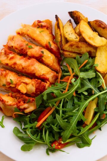 Close-up of sliced baked chicken parmesan topped with melted cheese and tomato sauce, served with golden roasted potato wedges and a fresh arugula salad with shredded carrots and red bell pepper.