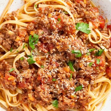 A close-up of hearty Italian ground beef linguine served in a white bowl, topped with a rich tomato meat sauce, fresh parsley, and a sprinkle of Parmesan cheese. The golden linguine is coated in the savory sauce with visible chunks of tomato and carrot, creating a rustic and comforting Italian pasta dish.
