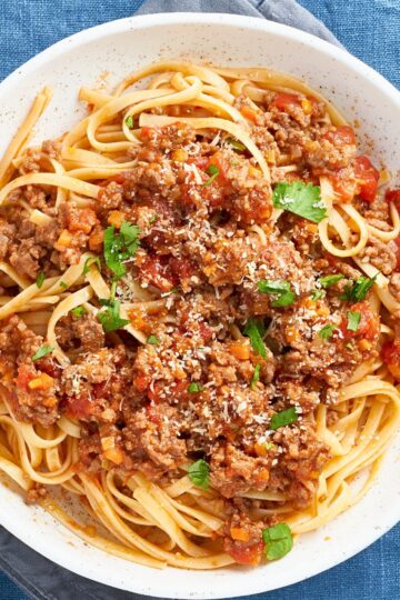 A close-up of hearty Italian ground beef linguine served in a white bowl, topped with a rich tomato meat sauce, fresh parsley, and a sprinkle of Parmesan cheese. The golden linguine is coated in the savory sauce with visible chunks of tomato and carrot, creating a rustic and comforting Italian pasta dish.