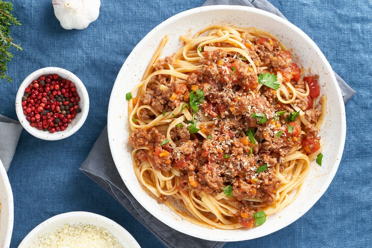 A white bowl filled with hearty Italian ground beef linguine, topped with a rich tomato and meat sauce, fresh parsley, and a sprinkle of Parmesan cheese. The dish sits on a blue tablecloth with a small bowl of red and black peppercorns, a bowl of grated Parmesan, and a garlic bulb nearby, creating a rustic Italian dinner setting.