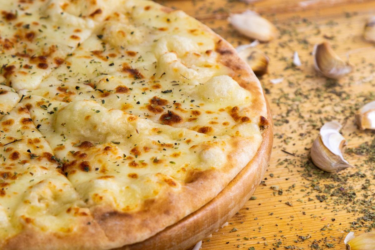 Close-up of a sliced pita pizza with bubbly melted mozzarella and dried herbs on a wooden board, with garlic cloves and oregano scattered around. Easy 10 minute pita pizza, quick weeknight dinner, garlic cheese flatbread.