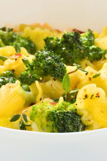 Close-up of baked broccoli macaroni and cheese in a white casserole dish: ruffled pasta coated in gooey cheddar, roasted broccoli florets, fresh thyme, black pepper, and red pepper flakes. Comfort food dinner, vegetarian pasta bake.