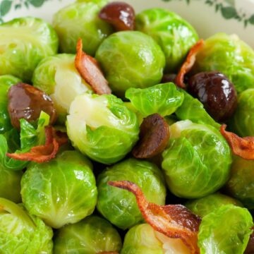 Close-up of bright green Brussels sprouts with roasted chestnuts and crisp bacon pieces in a cream bowl with a green vine rim; glossy, buttery finish. Christmas side dish recipe; Brussels sprouts with chestnuts and bacon.