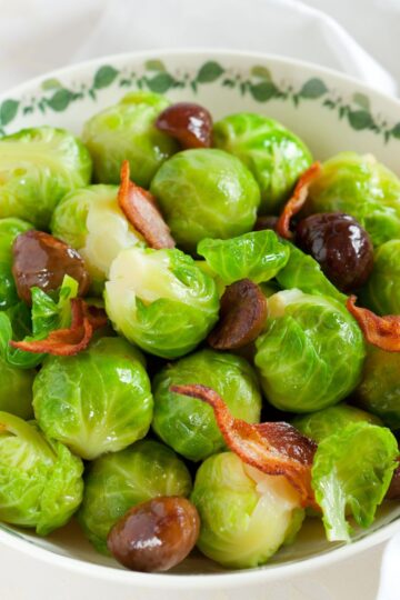 Close-up of bright green Brussels sprouts with roasted chestnuts and crisp bacon pieces in a cream bowl with a green vine rim; glossy, buttery finish. Christmas side dish recipe; Brussels sprouts with chestnuts and bacon.