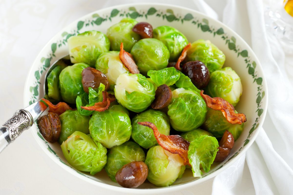 Bowl with vine-rim pattern filled with Brussels sprouts, roasted chestnuts, and crisp bacon; vintage silver spoon on the left and white linen backdrop. Holiday side dish; Brussels sprouts with chestnuts and bacon recipe.