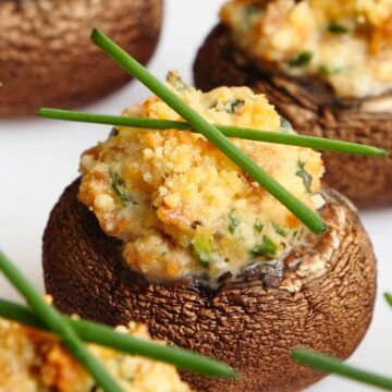 Close-up of baked stuffed mushrooms on a white plate, filled with creamy garlic-Parmesan cheese and herbs, golden and crumbly on top, garnished with fresh chives. Appetizer recipe; garlic parmesan stuffed mushrooms; party snack; holiday appetizer.