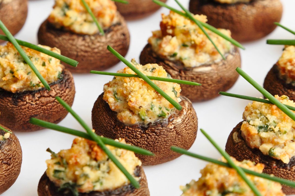 Bite-size stuffed mushrooms arranged on a white surface, topped with creamy garlic-Parmesan filling and herbs, garnished with fresh chive sticks and golden, crispy crumbs. Party appetizer recipe; garlic parmesan stuffed mushrooms.