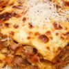 Close-up slice of crockpot lazy lasagna on a beige plate—golden, bubbly cheese topped with shredded Parmesan over layered noodles, beef, and chunky tomato sauce. Slow cooker pasta, easy dinner, Italian comfort food.