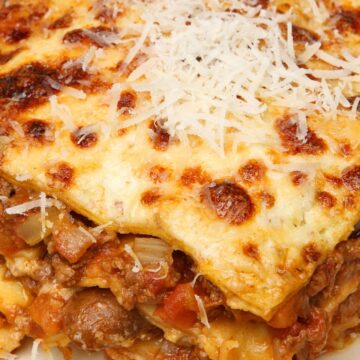Close-up slice of crockpot lazy lasagna on a beige plate—golden, bubbly cheese topped with shredded Parmesan over layered noodles, beef, and chunky tomato sauce. Slow cooker pasta, easy dinner, Italian comfort food.