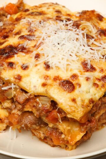 Close-up slice of crockpot lazy lasagna on a beige plate—golden, bubbly cheese topped with shredded Parmesan over layered noodles, beef, and chunky tomato sauce. Slow cooker pasta, easy dinner, Italian comfort food.