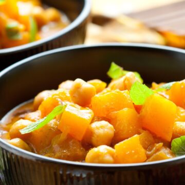 Close-up of sweet potato curry with chickpeas in a black bowl, garnished with small spinach leaves; a second bowl softly blurred behind on a bamboo mat in warm light.