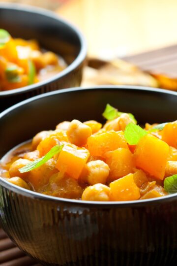 Close-up of sweet potato curry with chickpeas in a black bowl, garnished with small spinach leaves; a second bowl softly blurred behind on a bamboo mat in warm light.