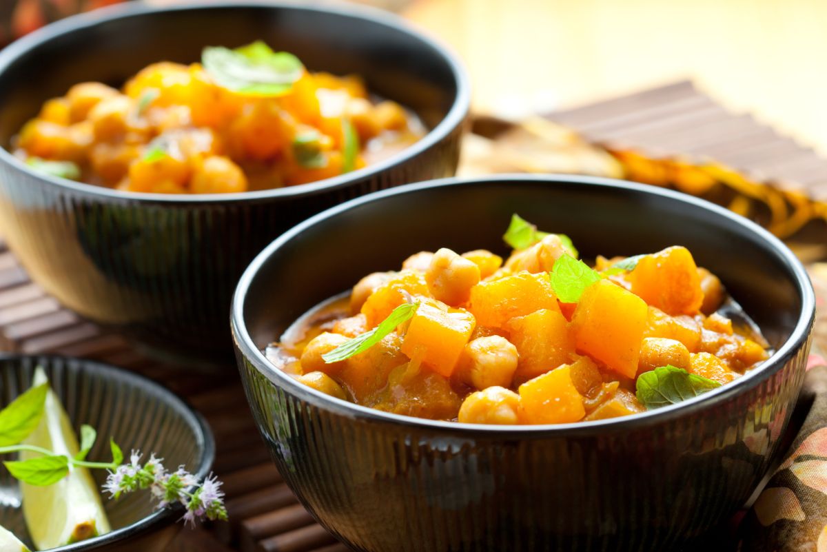 Two black bowls of sweet potato and chickpea curry, topped with small green spinach/herb leaves, set on a bamboo mat in warm light. Chunky orange cubes in a thick sauce; second bowl softly blurred behind.