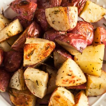 Close-up of roasted red potatoes with rosemary, quartered and golden with crispy edges in a white bowl on a rustic wood table. Coarse salt and herbs visible. Keywords: roasted red potatoes, rosemary potatoes, easy side dish, oven roasted recipe.