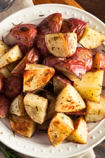 Close-up of roasted red potatoes with rosemary, quartered and golden with crispy edges in a white bowl on a rustic wood table. Coarse salt and herbs visible. Keywords: roasted red potatoes, rosemary potatoes, easy side dish, oven roasted recipe.