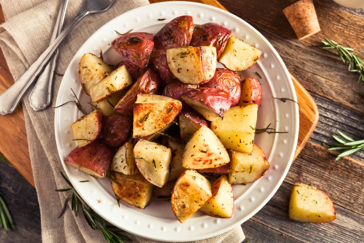 Bowl of golden roasted red potatoes with rosemary on a white plate, quartered and crispy-edged, set on a rustic wood table with linen napkin and silverware. Fresh rosemary sprigs around the plate. Keywords: roasted red potatoes, rosemary potatoes, easy side dish, oven roasted recipe.