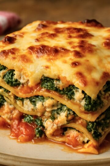 Close-up of ground chicken and spinach lasagna: golden, bubbly mozzarella on top, layered pasta with ricotta and spinach, and tomato sauce oozing onto a beige plate. Italian comfort food, high-protein chicken lasagna dinner recipe.