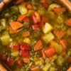 Close-up of a wooden bowl filled with rustic Italian vegetable soup—chunky carrots, potatoes, celery, red bell pepper, and herbs in a clear broth. Cozy, homemade, healthy soup recipe; Italian comfort food.