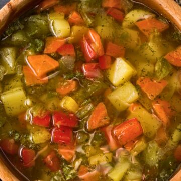 Close-up of a wooden bowl filled with rustic Italian vegetable soup—chunky carrots, potatoes, celery, red bell pepper, and herbs in a clear broth. Cozy, homemade, healthy soup recipe; Italian comfort food.
