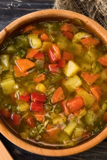 Close-up of a wooden bowl filled with rustic Italian vegetable soup—chunky carrots, potatoes, celery, red bell pepper, and herbs in a clear broth. Cozy, homemade, healthy soup recipe; Italian comfort food.