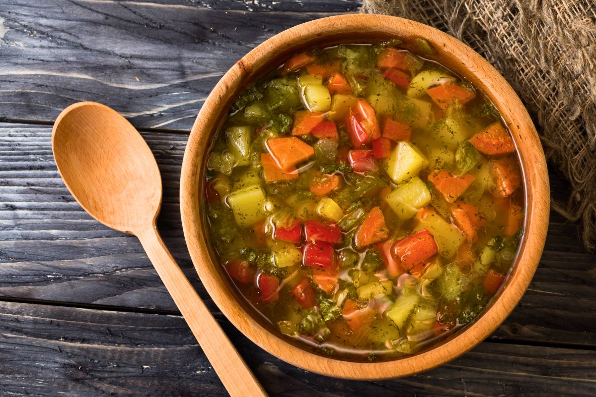 Overhead shot of a wooden bowl of rustic Italian vegetable soup on a dark wood table with a wooden spoon. Chunky carrots, potatoes, celery, red peppers, and herbs in a clear broth. Cozy, homemade, healthy soup recipe, Italian comfort food.