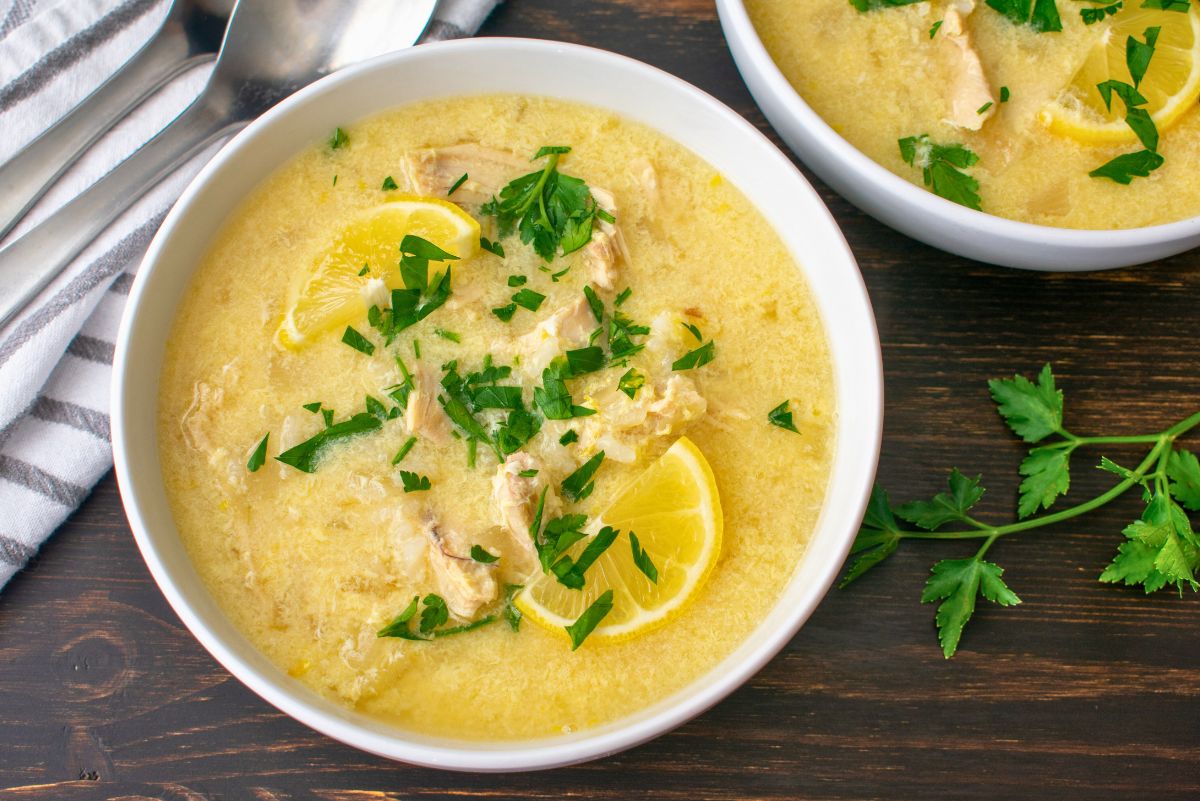 Greek Avgolemono chicken soup in white bowls on a dark wood table—creamy egg-lemon broth with shredded chicken, lemon wedges, and chopped parsley. A striped napkin and spoons sit at left, with fresh parsley sprigs on the table.