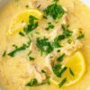 Close-up of Greek Avgolemono chicken soup—creamy egg-lemon broth with shredded chicken, lemon wedges, and chopped parsley in a white bowl on a dark wood table. Kitchen photography, comfort food, soup recipe.
