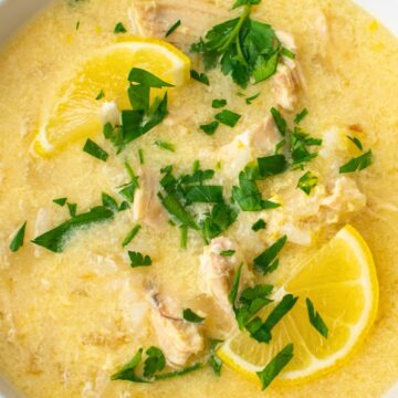 Close-up of Greek Avgolemono chicken soup—creamy egg-lemon broth with shredded chicken, lemon wedges, and chopped parsley in a white bowl on a dark wood table. Kitchen photography, comfort food, soup recipe.