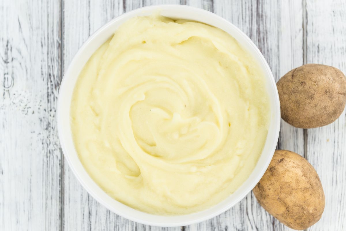Bowl of creamy cauliflower mashed potatoes with smooth swirls in a white dish on rustic white wood, two whole russet potatoes beside it.