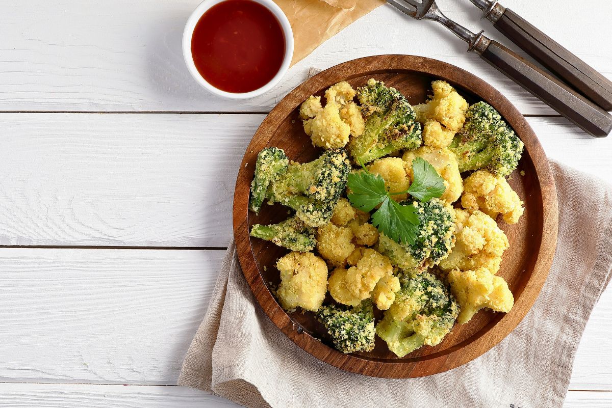 Baked broccoli and cauliflower florets coated in lemon herb crumbs and Parmesan, served in a wooden bowl with a parsley garnish. A small bowl of red dipping sauce sits nearby, with two forks and a beige napkin on a white wood table. Healthy vegetable side dish.